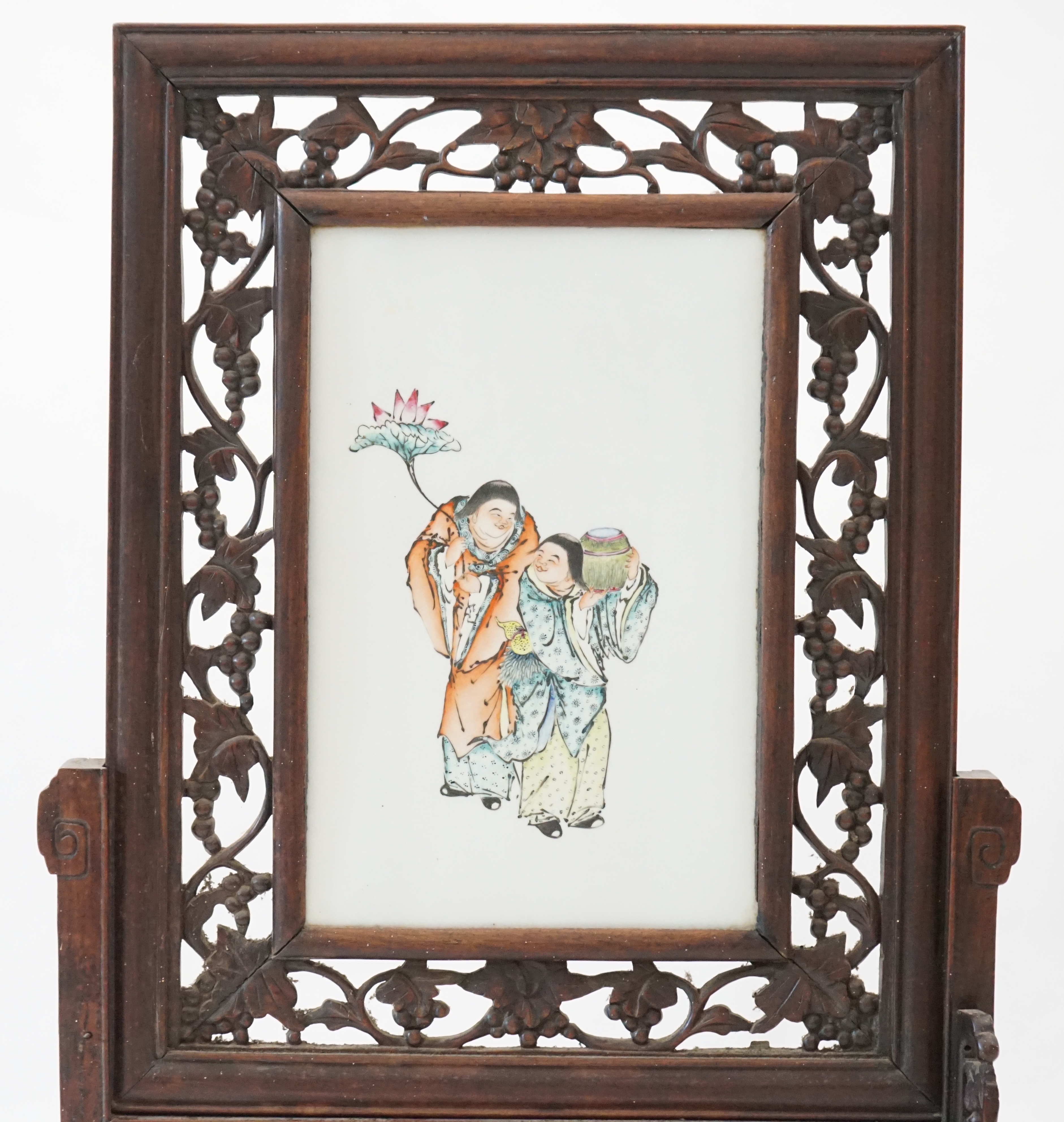 A Chinese enamelled porcelain and hardwood table screen, Republic period, painted with the HeHe Erxian, holding a sprig of lotus and a box and cover, the plaque 37.5cm x 24cm, the wood stand and frame carved with vines,
